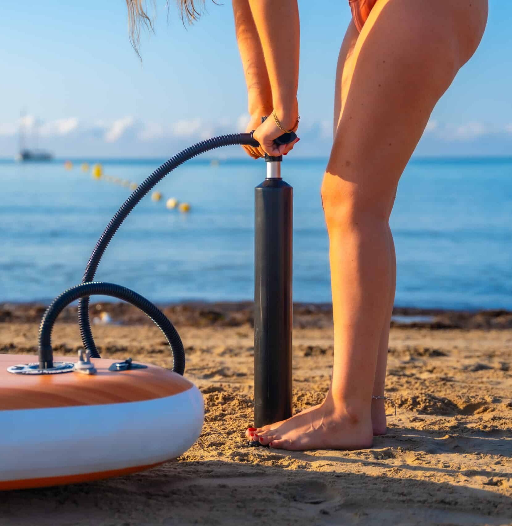 Woman inflating a paddleboard on a sandy beach, preparing for a summer paddle surfing session. Calm ocean and clear sky in the background